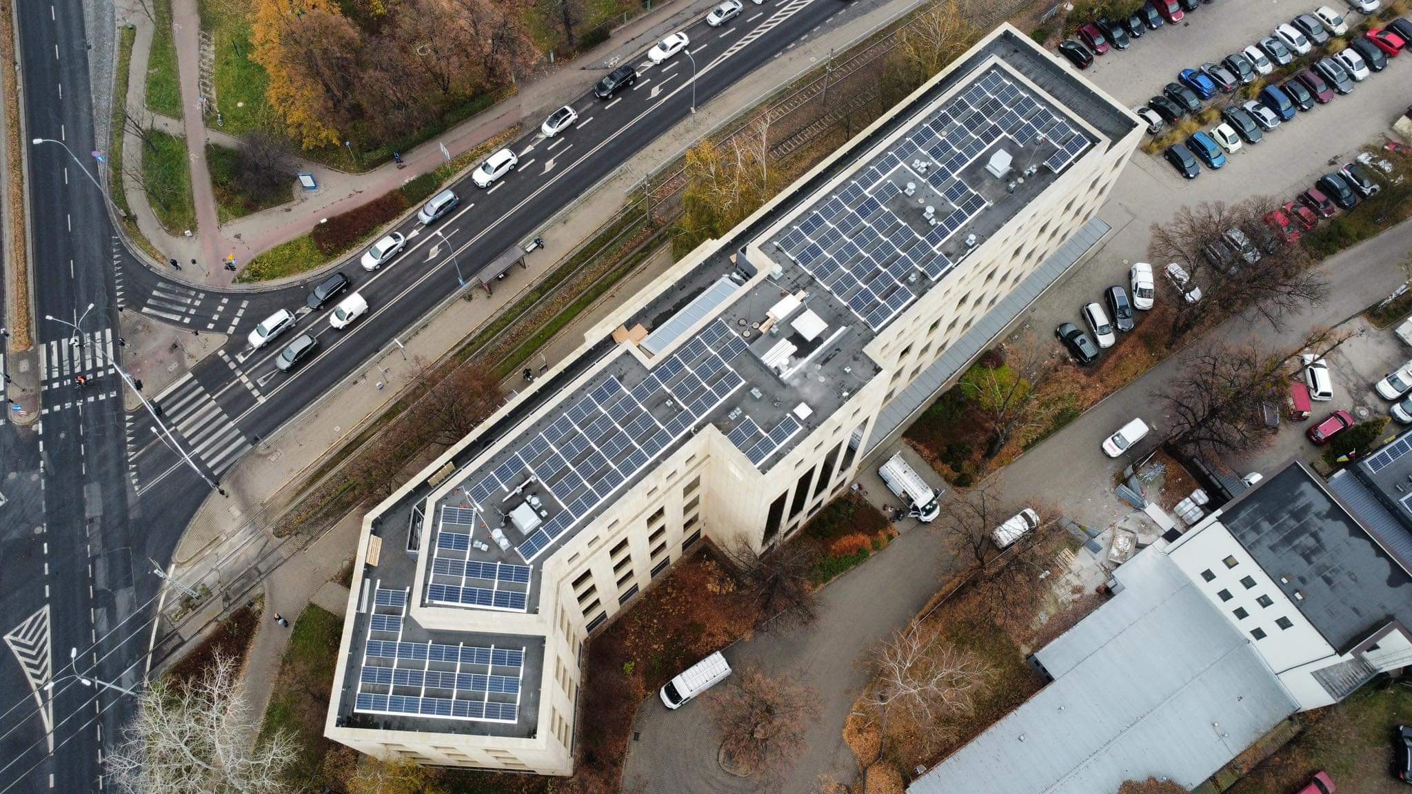 Widok z drona na budynek biurowy z panelami słonecznymi na dachu, obok ulica z ruchem samochodowym i parking.