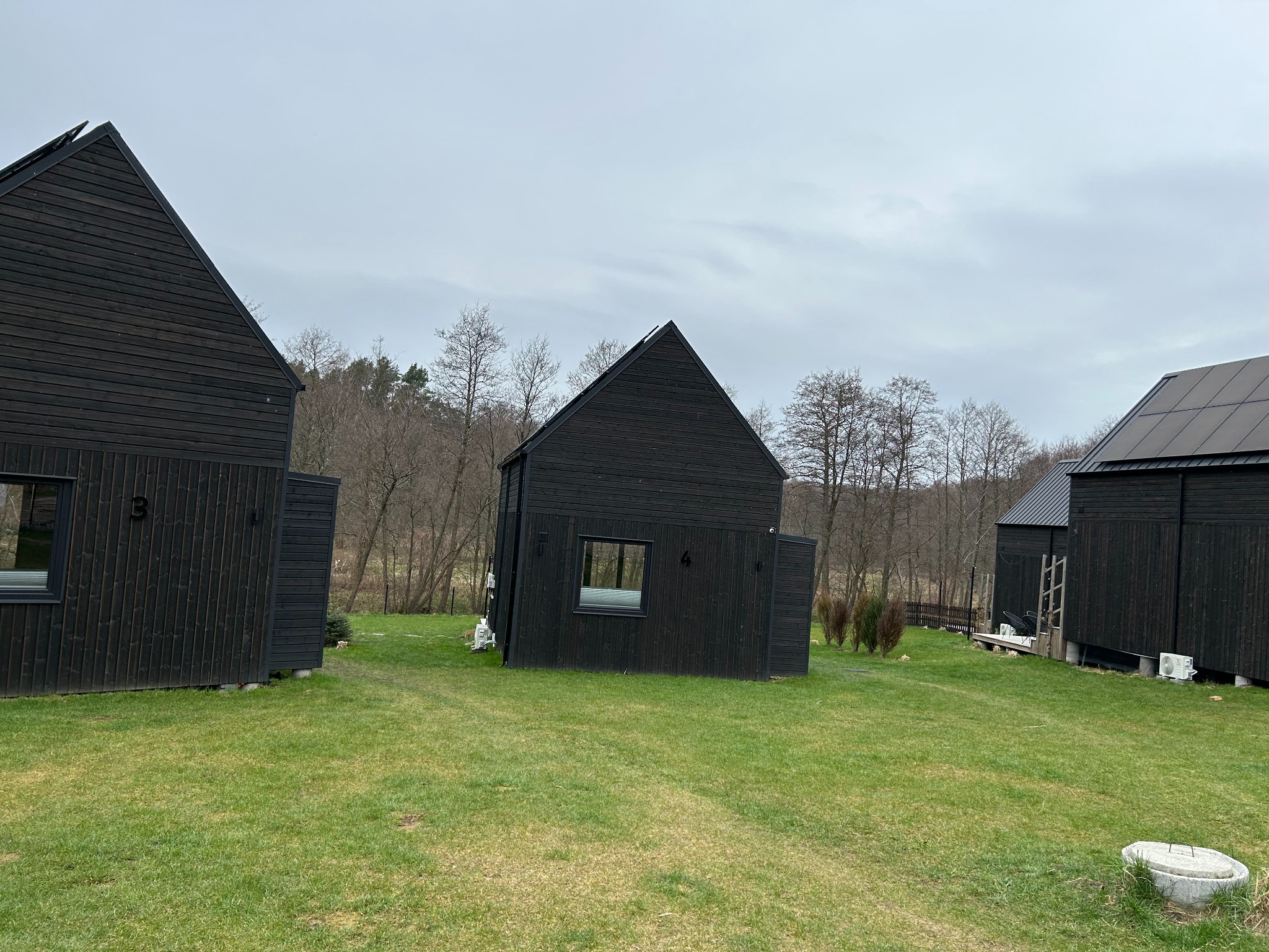 Trzy nowoczesne, czarne, drewniane domki letniskowe z panelami słonecznymi na dachu jednego z nich, stojące na zielonym trawniku w pochmurny dzień.