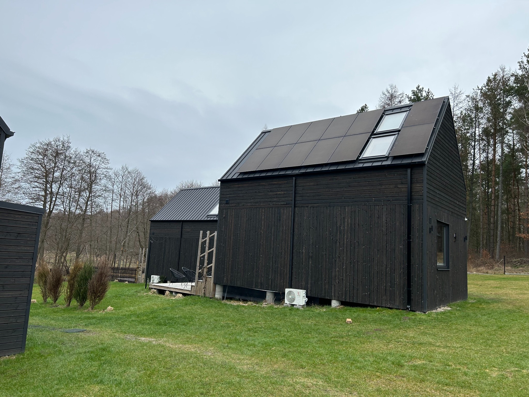 Nowoczesny, czarny dom z panelami fotowoltaicznymi i oknami dachowymi na dachu, otoczony zielonym trawnikiem i drzewami w tle.