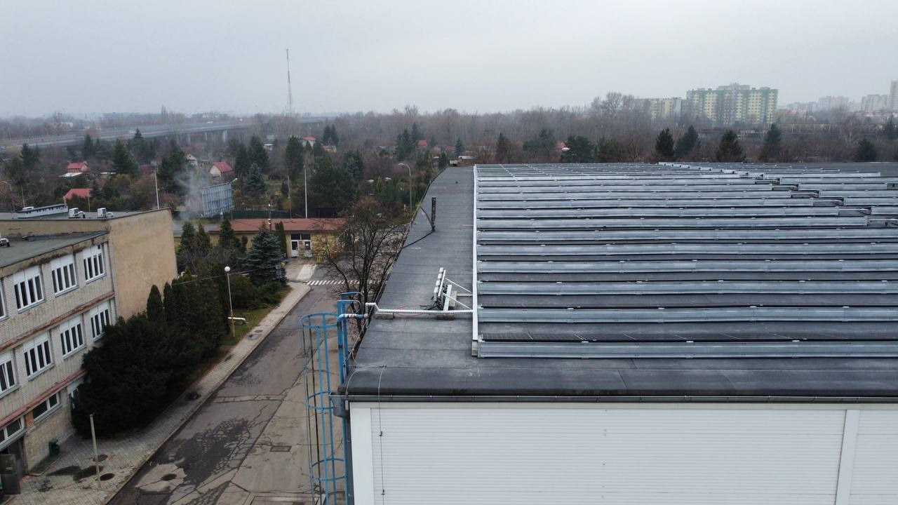 Szereg paneli słonecznych na płaskim dachu budynku przemysłowego w pochmurny dzień, z widoczną drabinką inspekcyjną i fragmentem otoczenia miejskiego w tle.