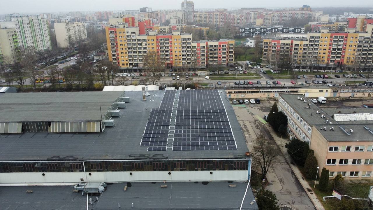 Widok z góry na dużą instalację fotowoltaiczną na płaskim dachu budynku przemysłowego w szarej miejskiej scenerii, z blokami mieszkalnymi w tle.