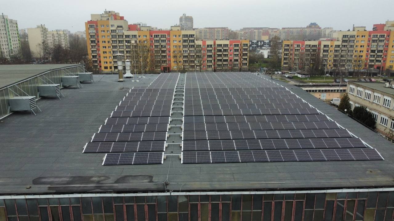 Rozległa instalacja paneli słonecznych na płaskim dachu budynku przemysłowego, widok z góry, na horyzoncie zabudowa miejska.