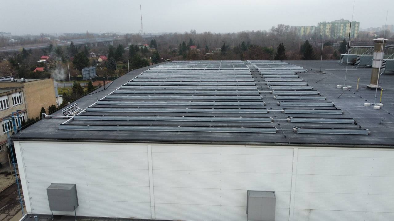 Rząd paneli na płaskim dachu budynku przemysłowego w pochmurny dzień, widok z góry, w tle zabudowa miejska i drzewa.