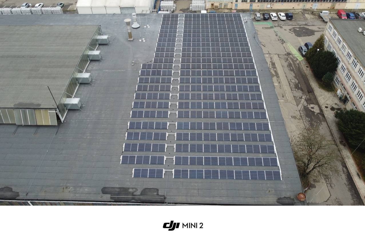 Widok z drona na rozległą instalację paneli słonecznych na płaskim dachu budynku przemysłowego, z widocznymi elementami wentylacji i fragmentem parkingu w tle.