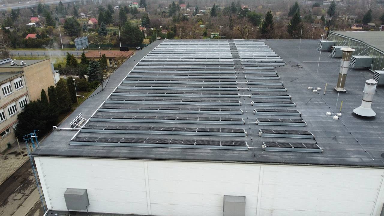 Widok z góry na dużą instalację fotowoltaiczną na płaskim dachu budynku przemysłowego z widocznymi elementami wentylacji i drabinką dostępową.
