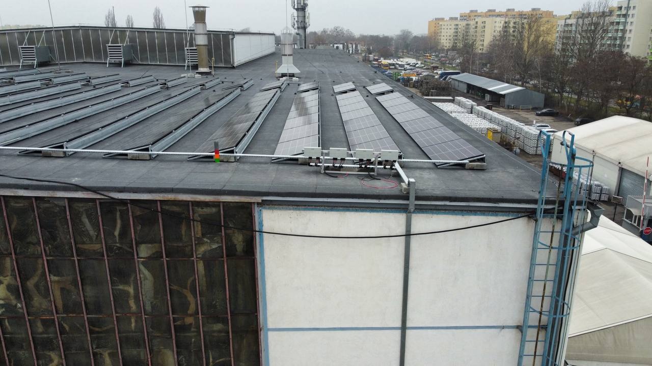 Instalacja paneli słonecznych na dachu budynku przemysłowego, widoczne rzędy paneli, konstrukcja wsporcza, falowniki, drabina na ścianie budynku, w tle zabudowa miejska.