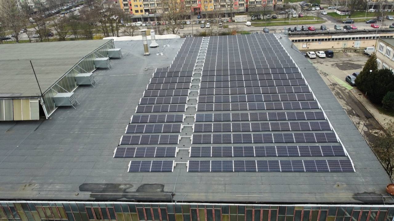 Widok z góry na dużą instalację paneli słonecznych na płaskim dachu budynku przemysłowego, z widocznymi elementami wentylacji i otaczającą zabudową miejską w tle.