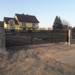 Nowoczesna, pozioma brama przesuwna w kolorze antracytowym, zamontowana na posesji z dwupiętrowym domem w tle, widok z zewnątrz na żwirową drogę dojazdową.