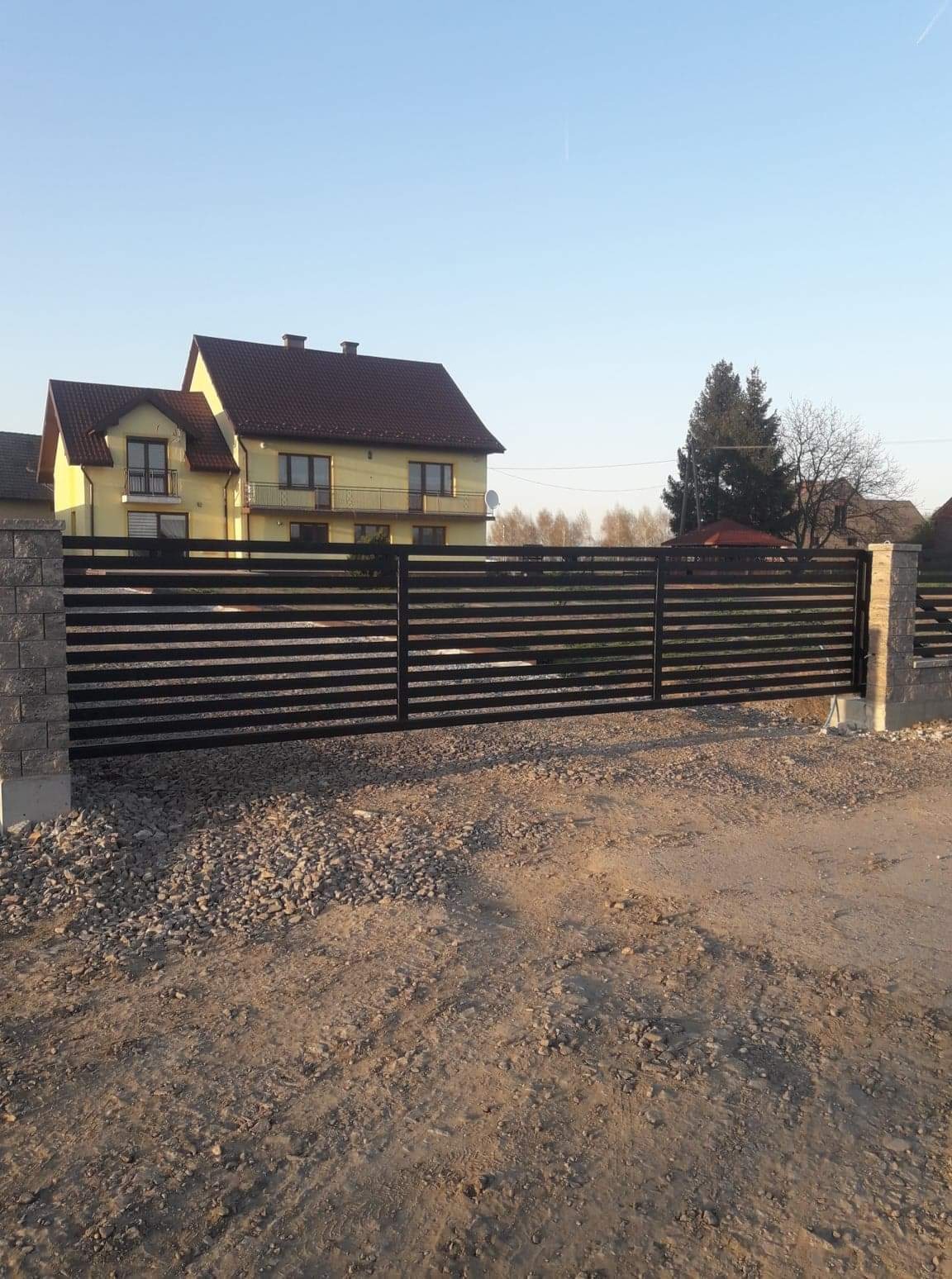 Nowoczesna, pozioma brama przesuwna w kolorze antracytowym, zamontowana na posesji z dwupiętrowym domem w tle, widok z zewnątrz na żwirową drogę dojazdową.