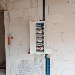 El-ton - Otwarta skrzynka rozdzielcza z widocznymi bezpiecznikami i przewodami na ścianie z betonu komórkowego, zasilanie doprowadzone w peszlach.