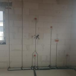 El-ton - Surowe wnętrze w trakcie prac: pionowe metalowe rurki i poziome czarne kable elektryczne prowadzone po ścianie z białych bloczków, okrągłe pomarańczowe puszki instalacyjne w ścianie, okno...