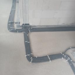 El-ton - Szereg szarych i białych rurek instalacyjnych, przymocowanych do szarej ściany w surowym stanie, układających się w równoległe linie i zakręty.