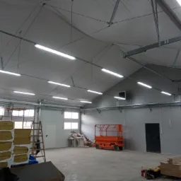 Wnętrze hali magazynowej z zamontowanym oświetleniem liniowym LED, widoczna podnośnik koszowy, materiały izolacyjne i drabina.