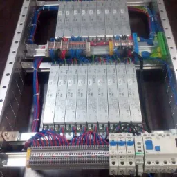 Rozdzielnica elektryczna w metalowej obudowie z licznymi przewodami, zabezpieczeniami i modułami, widoczne złącza śrubowe i przewody w kolorach niebieskim, czerwonym i zielonym.