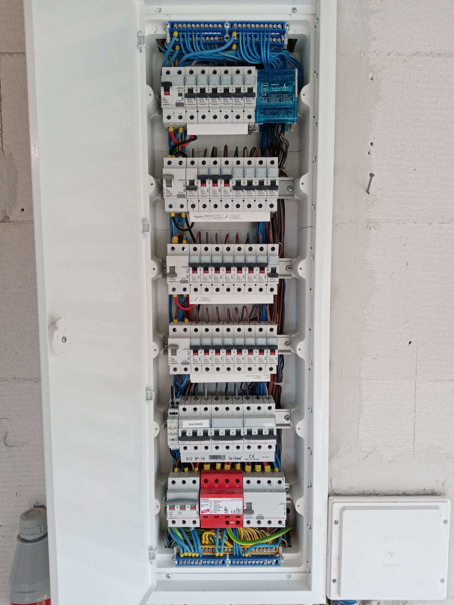 Otwarta skrzynka rozdzielcza z zabezpieczeniami różnicowoprądowymi i nadprądowymi Schneider Electric i Legrand, widoczne przewody w kolorach niebieskim, żółto-zielonym, brązowym i czarnym...
