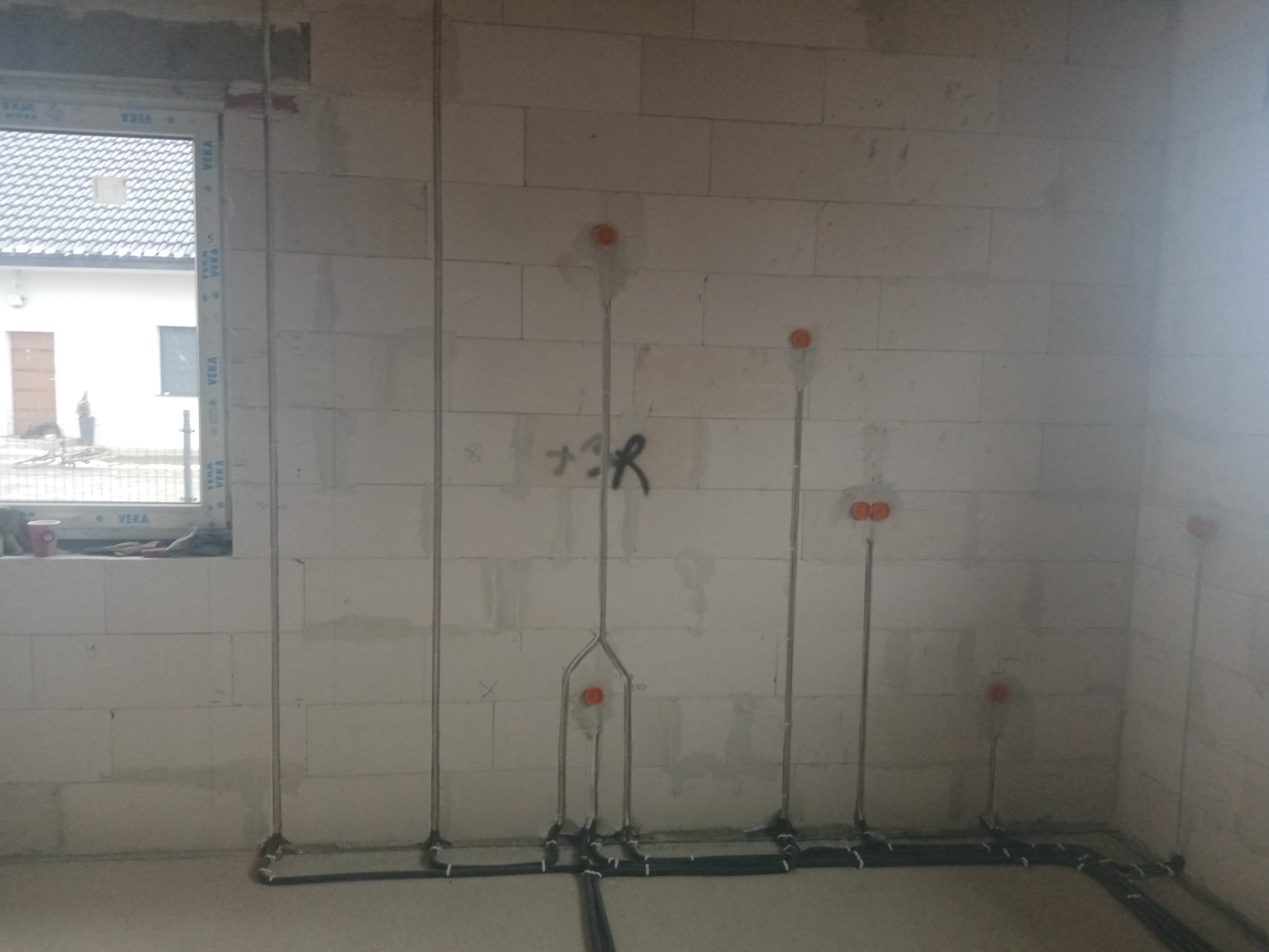 Surowe wnętrze w trakcie prac: pionowe metalowe rurki i poziome czarne kable elektryczne prowadzone po ścianie z białych bloczków, okrągłe pomarańczowe puszki instalacyjne w ścianie, okno...