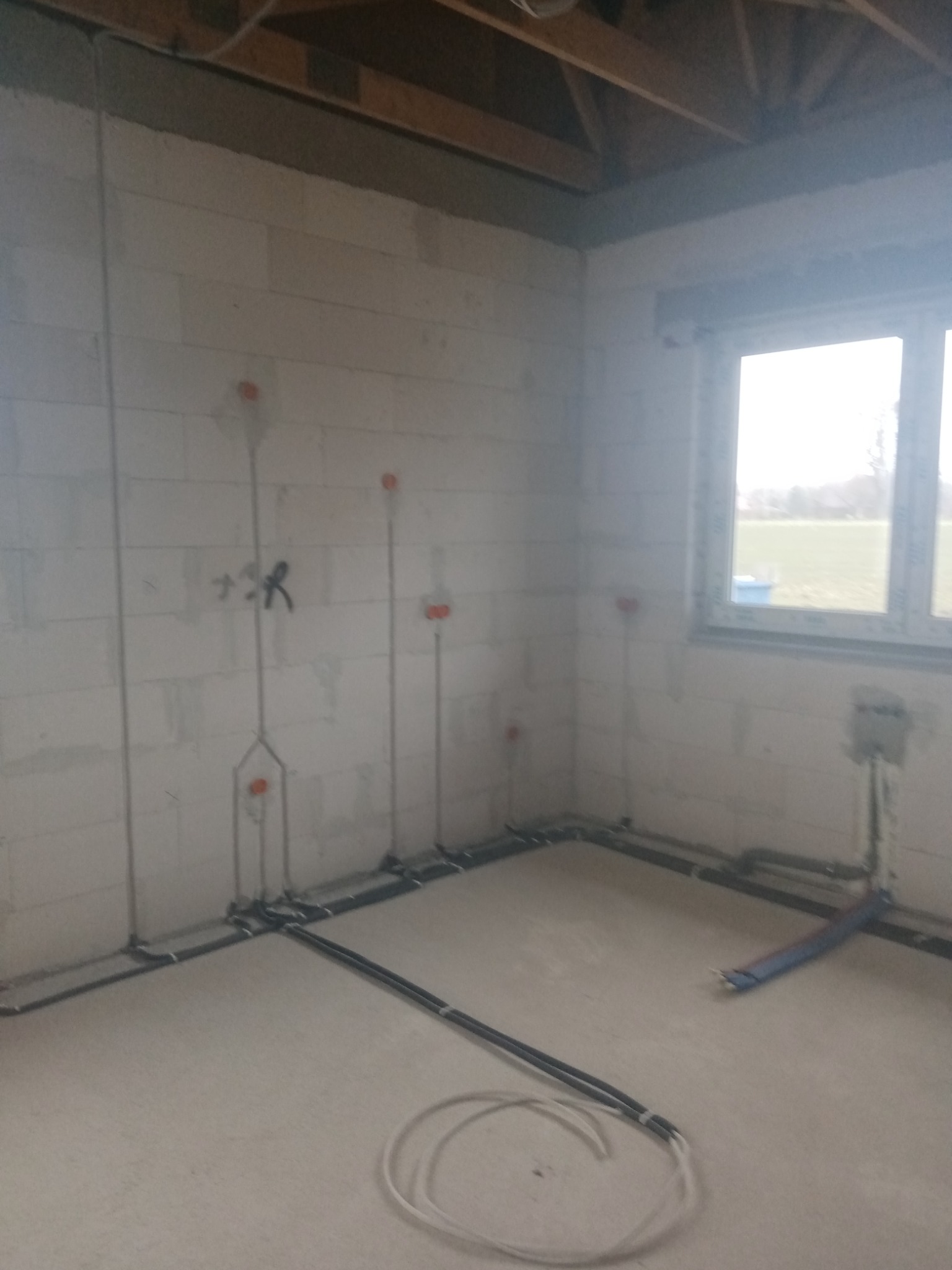 Wnętrze w trakcie budowy z widocznymi przewodami elektrycznymi poprowadzonymi po ścianach z bloczków i podłodze, puszki instalacyjne w ścianach, widok na okno z zewnątrz.