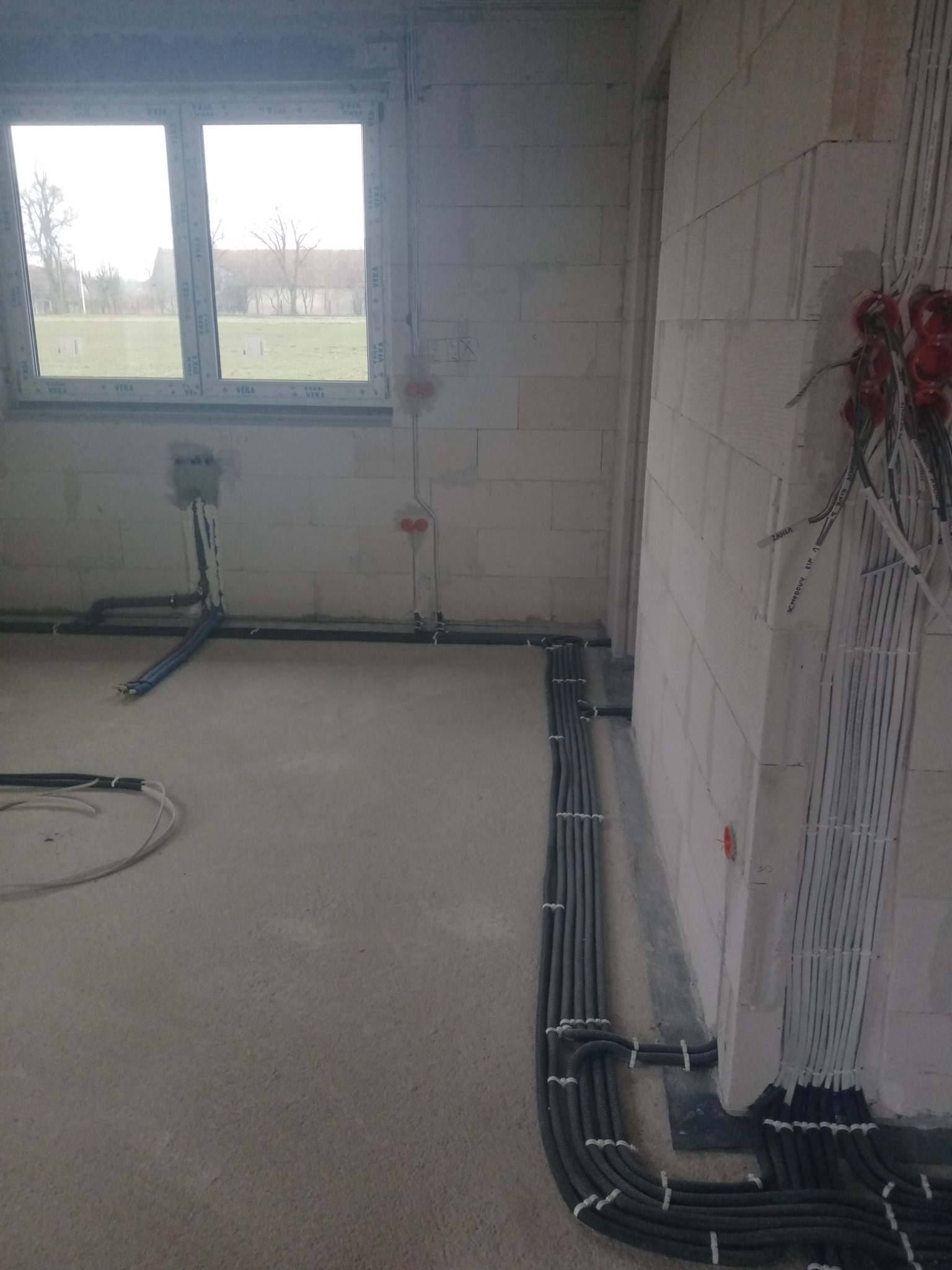 Surowy stan wnętrza z widocznymi przewodami elektrycznymi w peszlach, poprowadzonymi po ścianach i podłodze, oraz puszkami instalacyjnymi w ścianach z betonu komórkowego. Widoczne okno z widokiem...