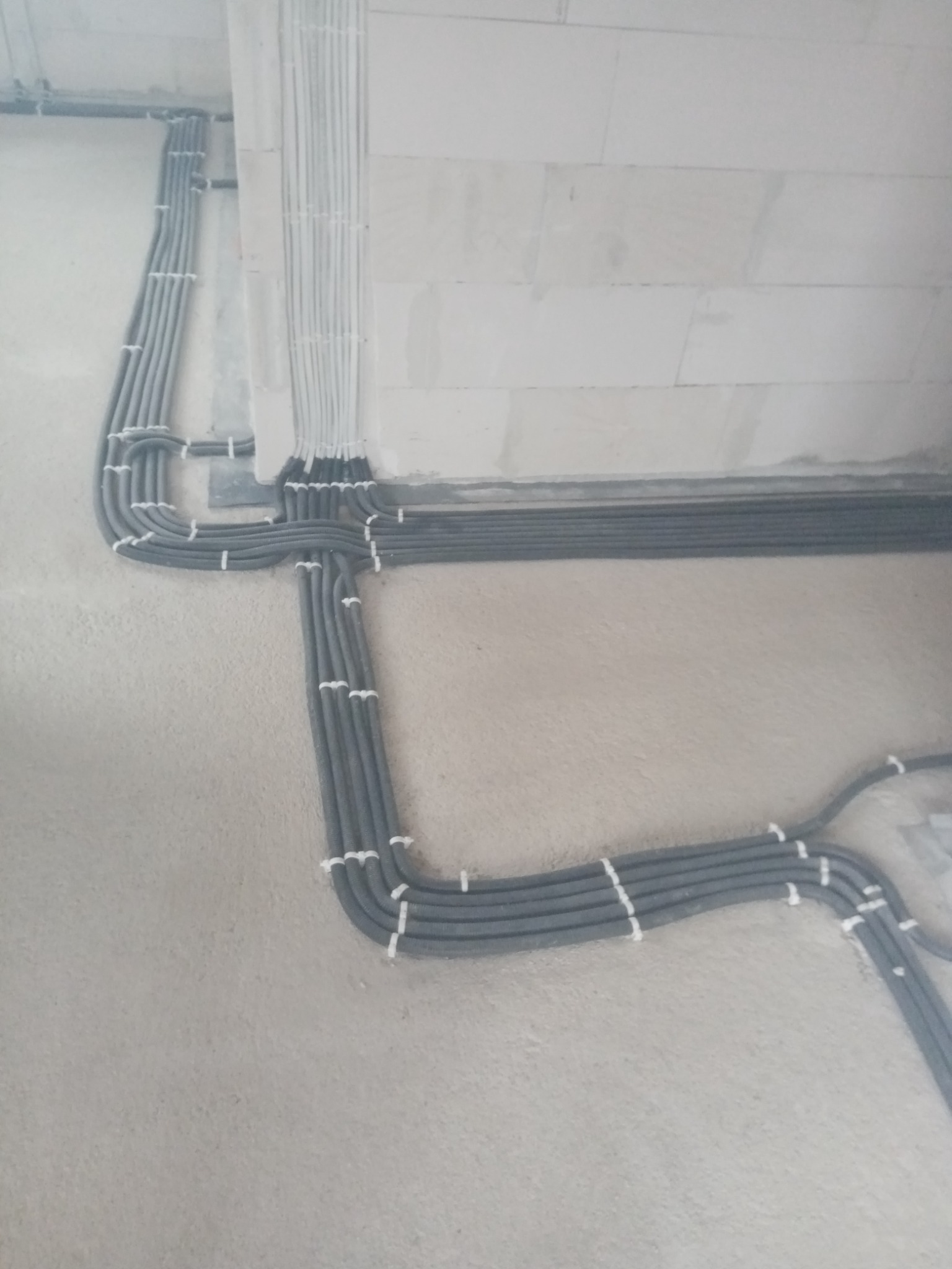 Szereg szarych i białych rurek instalacyjnych, przymocowanych do szarej ściany w surowym stanie, układających się w równoległe linie i zakręty.