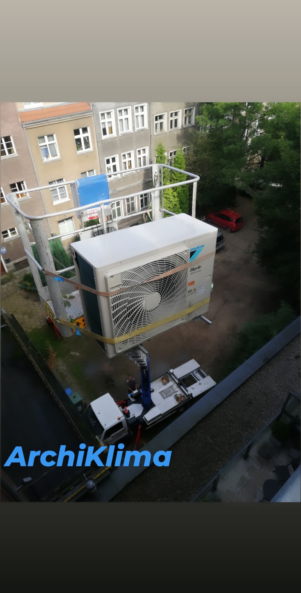 Montaż zewnętrznej jednostki klimatyzacji Daikin SkyAir za pomocą podnośnika, widok z góry na budynek mieszkalny i podwórko.