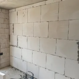 Ściana z białych bloczków połączonych zaprawą, z widocznym miejscami ubytkiem zaprawy, na podłodze leży wiertarka udarowa.