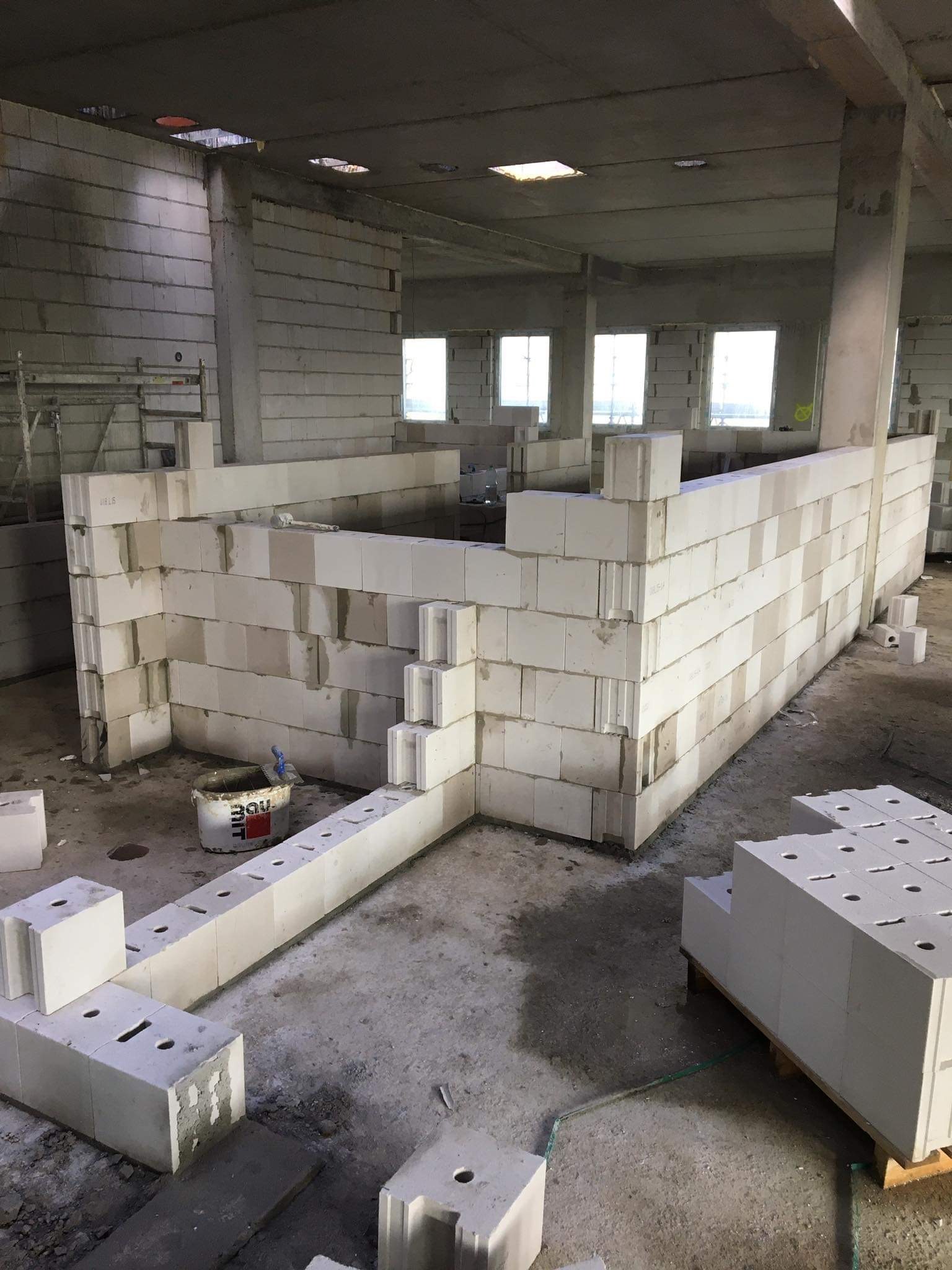Wnętrze budynku w trakcie budowy, ściany z białych bloczków z betonu komórkowego, widoczny kubeł z zaprawą i kielnia, surowy betonowy strop z otworami okiennymi, podłoże w trakcie prac murarskich.