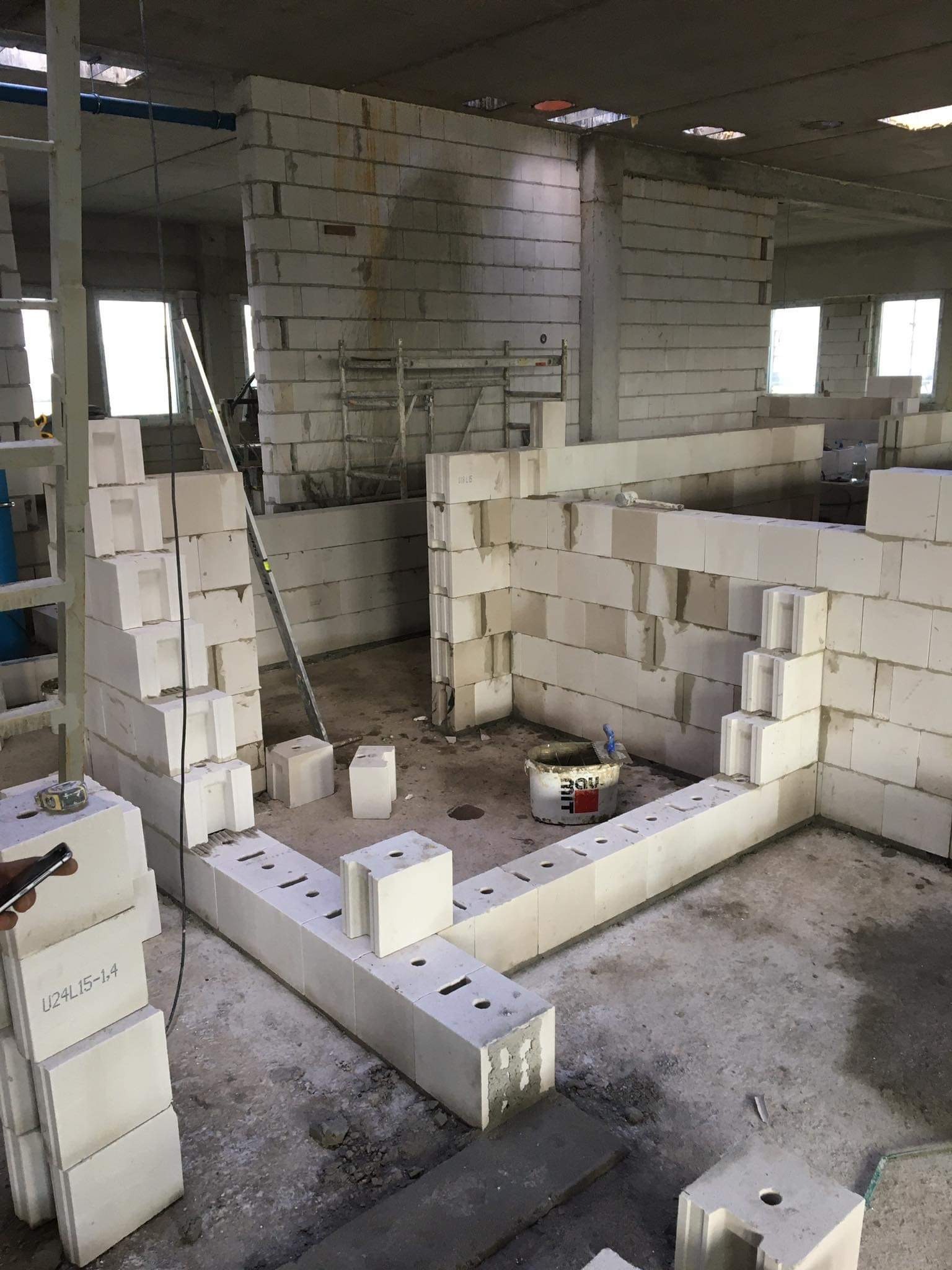 Wnętrze w trakcie budowy ścian z bloczków betonu komórkowego; widoczny częściowo wzniesiony mur, stosy bloczków z oznaczeniami U24L15-1.4, wiadro z zaprawą i poziomica oparta o ścianę.