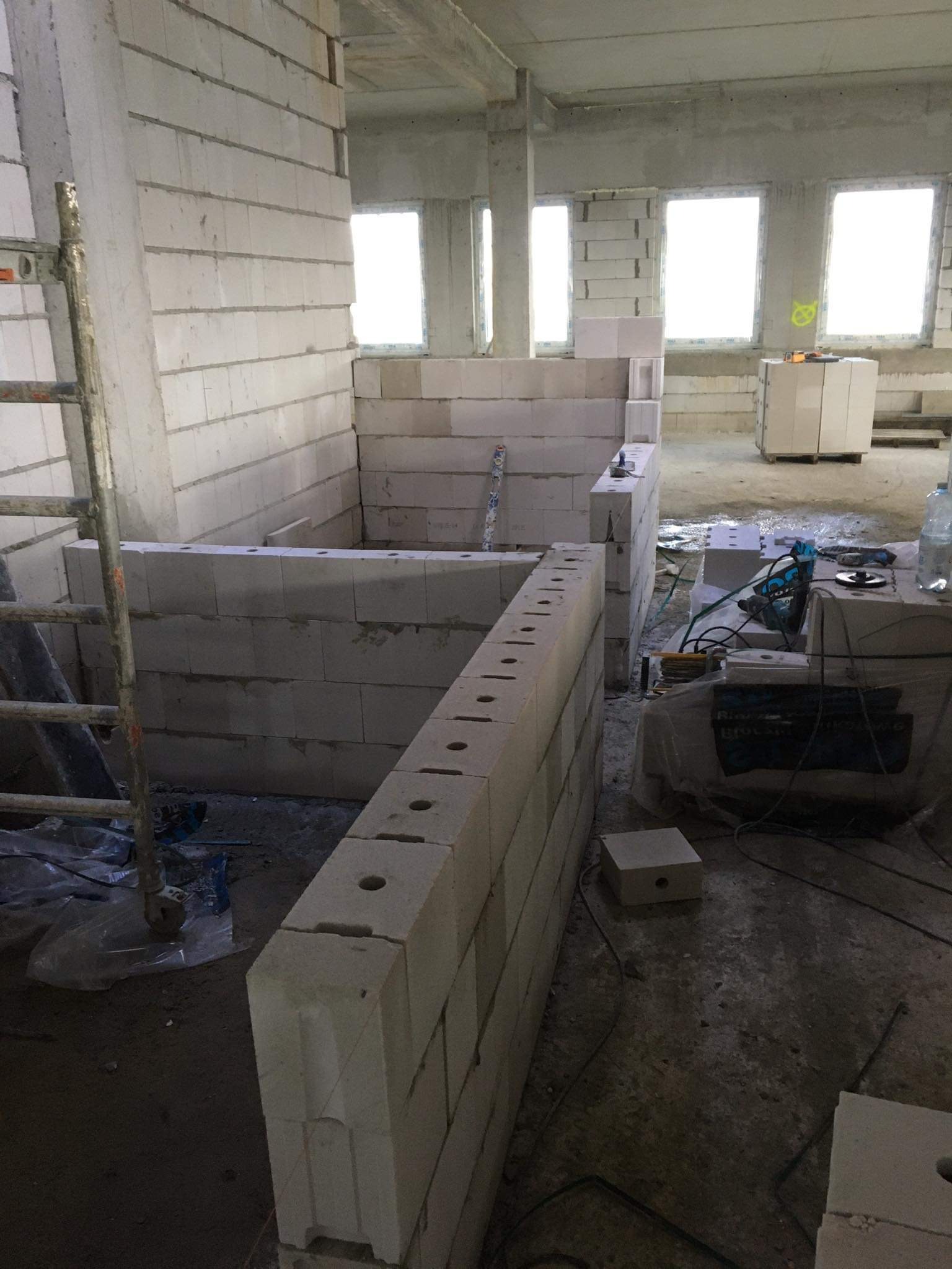 Wnętrze w trakcie budowy z widocznymi ścianami z bloczków, narzędziami i butelką wody, ukazujące etap prac murarskich przed tynkowaniem.