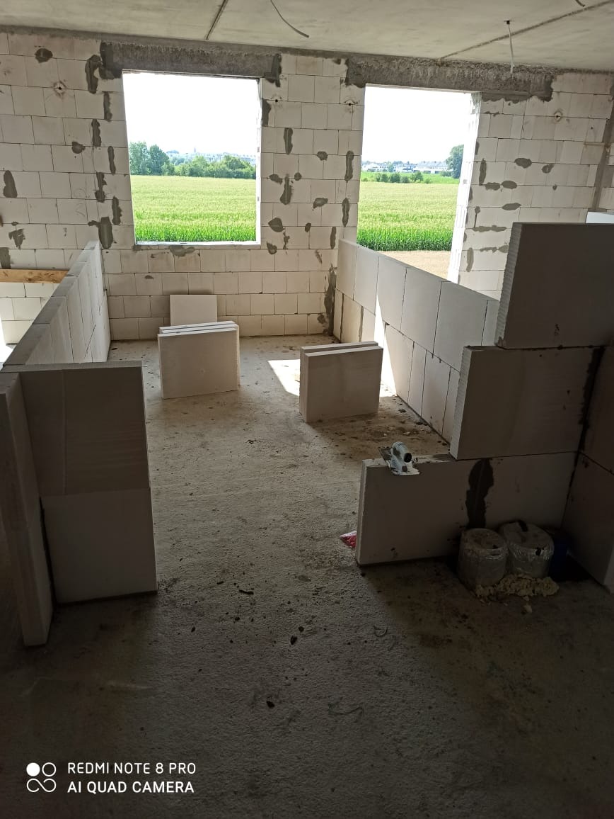 Wnętrze budynku w trakcie murowania ścian z bloczków, widoczne otwory okienne z widokiem na zielone pola, surowy beton na podłodze, ślady zaprawy murarskiej na ścianach.