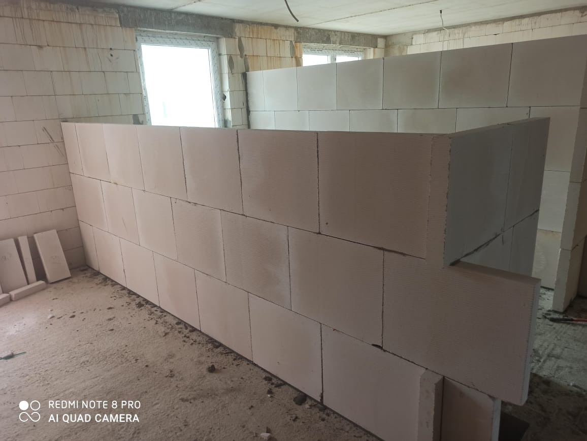 Wnętrze w trakcie budowy, ściana z białych bloczków z widocznymi spoinami, surowy betonowy sufit, okno w tle, przygotowane bloczki na podłodze, widok z niskiej perspektywy.
