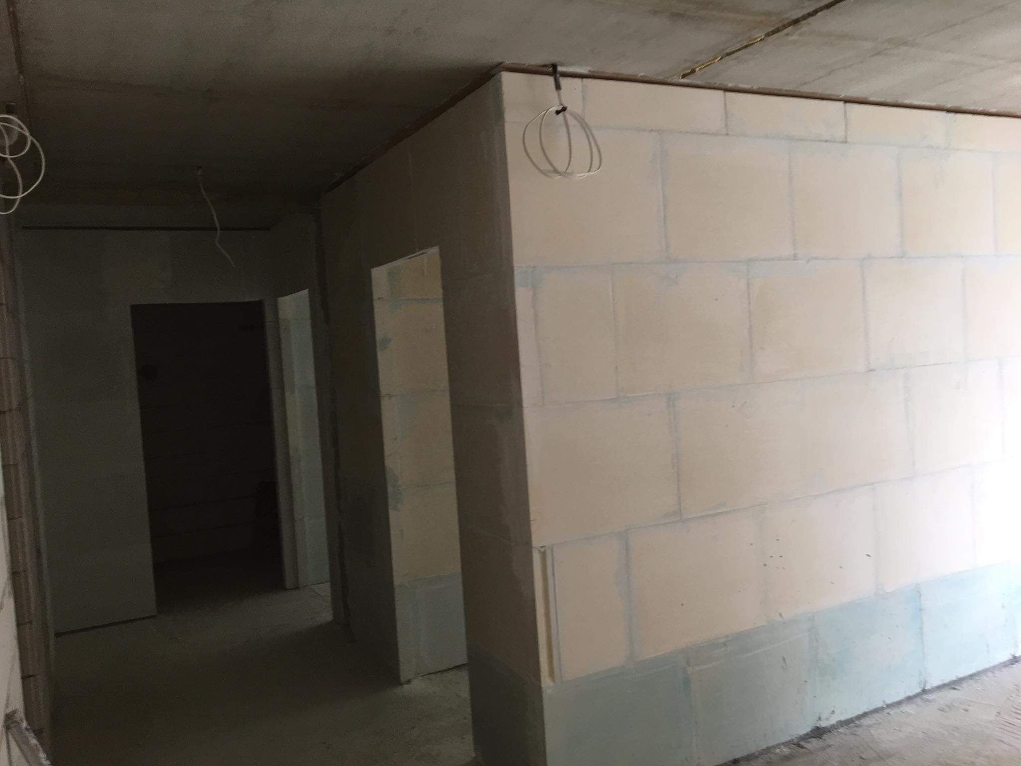 Wnętrze w stanie deweloperskim: ściany z betonu komórkowego częściowo pokryte szarą warstwą hydroizolacji, widoczne kable elektryczne zwisające z sufitu.
