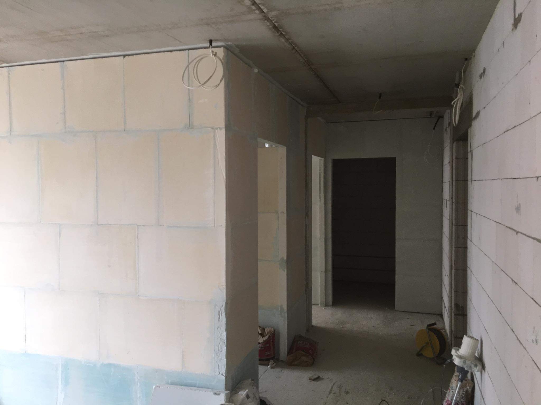 Wnętrze w stanie surowym z widocznymi ścianami z bloczków, częściowo przygotowanymi pod tynkowanie maszynowe, z widocznymi kablami i workami z materiałami budowlanymi.