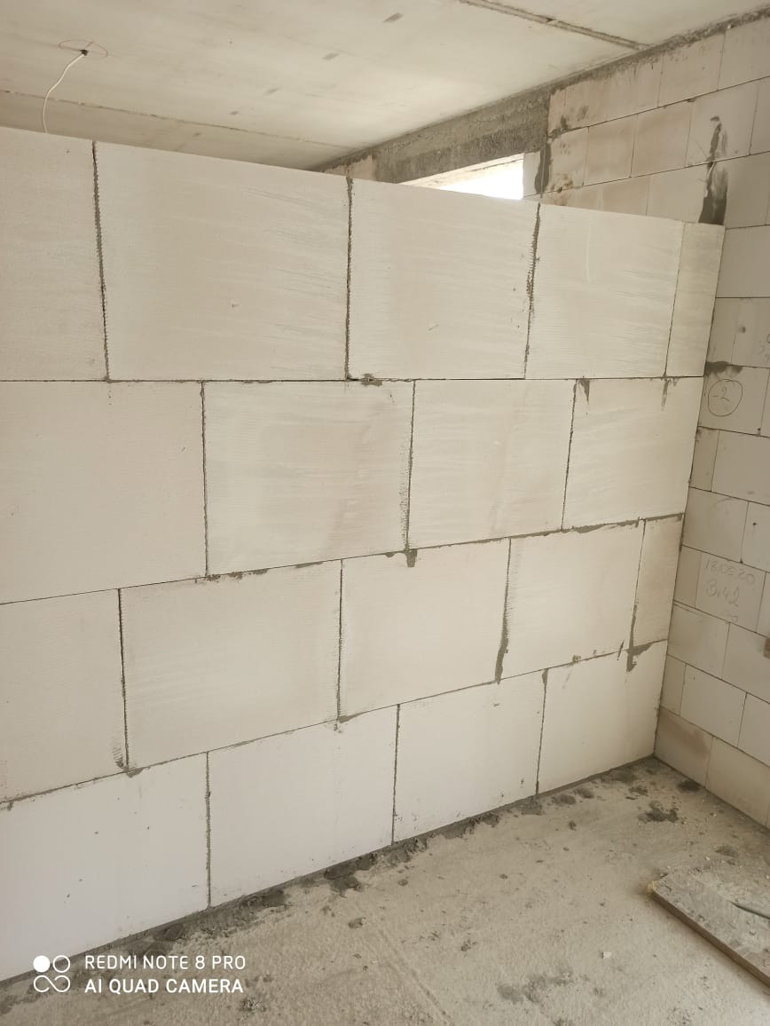 Ściana z białych bloczków z betonu komórkowego, świeżo wymurowana w surowym wnętrzu, widoczne ślady zaprawy, zarys okna w tle.