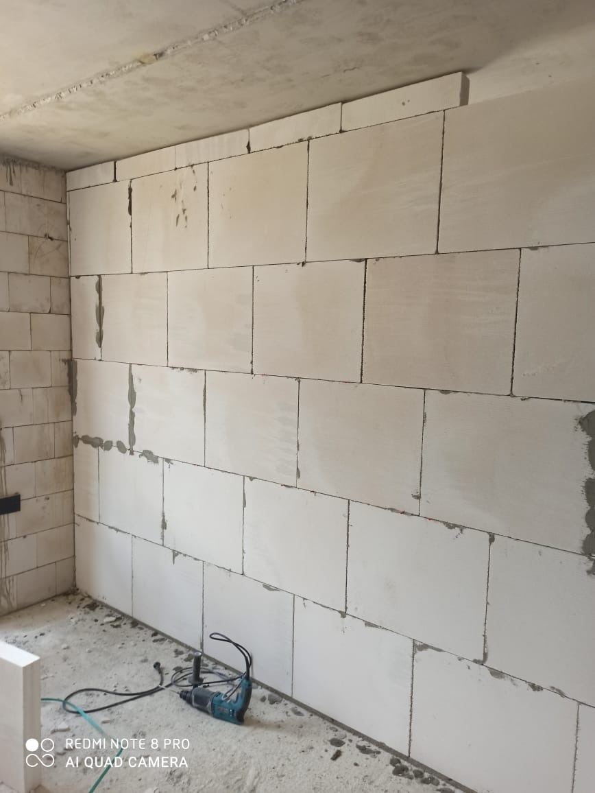 Ściana z białych bloczków połączonych zaprawą, z widocznym miejscami ubytkiem zaprawy, na podłodze leży wiertarka udarowa.