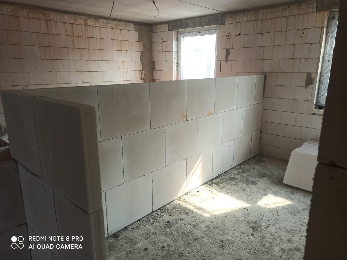 Wnętrze surowego pomieszczenia z wymurowanymi ścianami z białych bloczków, widoczne ślady po zalaniu na ścianach, okno w tle, podłoga betonowa.