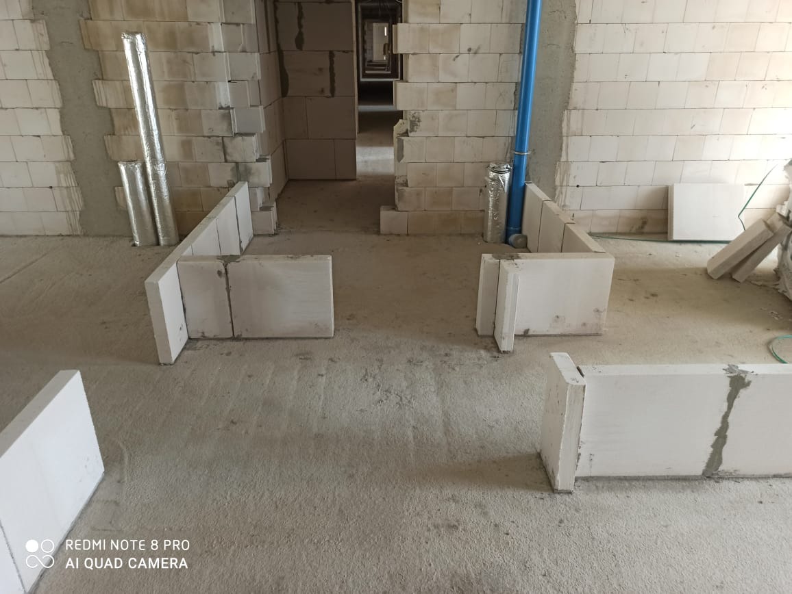 Wnętrze w trakcie budowy: ściany z białych bloczków, widoczne rury instalacyjne, surowy beton na podłodze, perspektywa w głąb pomieszczenia.