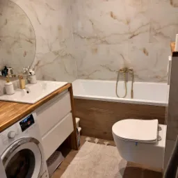 Nowoczesna łazienka z marmurowymi płytkami, drewnianymi akcentami i złotą armaturą, pralka z lewej strony.