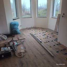 Firma ogólnobudowlana Damian Krzyżyk - Układanie paneli podłogowych w pokoju z trzema oknami, widoczne narzędzia: poziomica, maszyna do cięcia paneli, niebieski worek na śmieci, kliny poziomujące.
