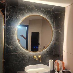 Firma ogólnobudowlana Damian Krzyżyk - Łazienka z umywalką, okrągłym lustrem z podświetleniem LED, baterią w kolorze złotym i czarną marmurową ścianą z widocznym odbiciem osoby w lustrze