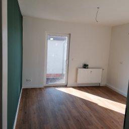 Firma ogólnobudowlana Damian Krzyżyk - Pusty pokój po remoncie z panelami podłogowymi, pomalowaną na zielono ścianą i grzejnikiem pod oknem balkonowym, widoczne wystające kable ze ściany.