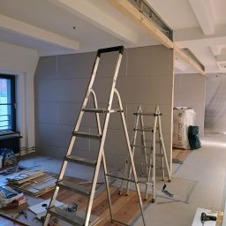 Firma ogólnobudowlana Damian Krzyżyk - Wnętrze w trakcie prac wykończeniowych: zamontowane płyty gipsowo-kartonowe na ścianach, widoczne dwie drabiny aluminiowe, narzędzia pracy i materiały budowlane na podłodze zabezpieczonej folią...