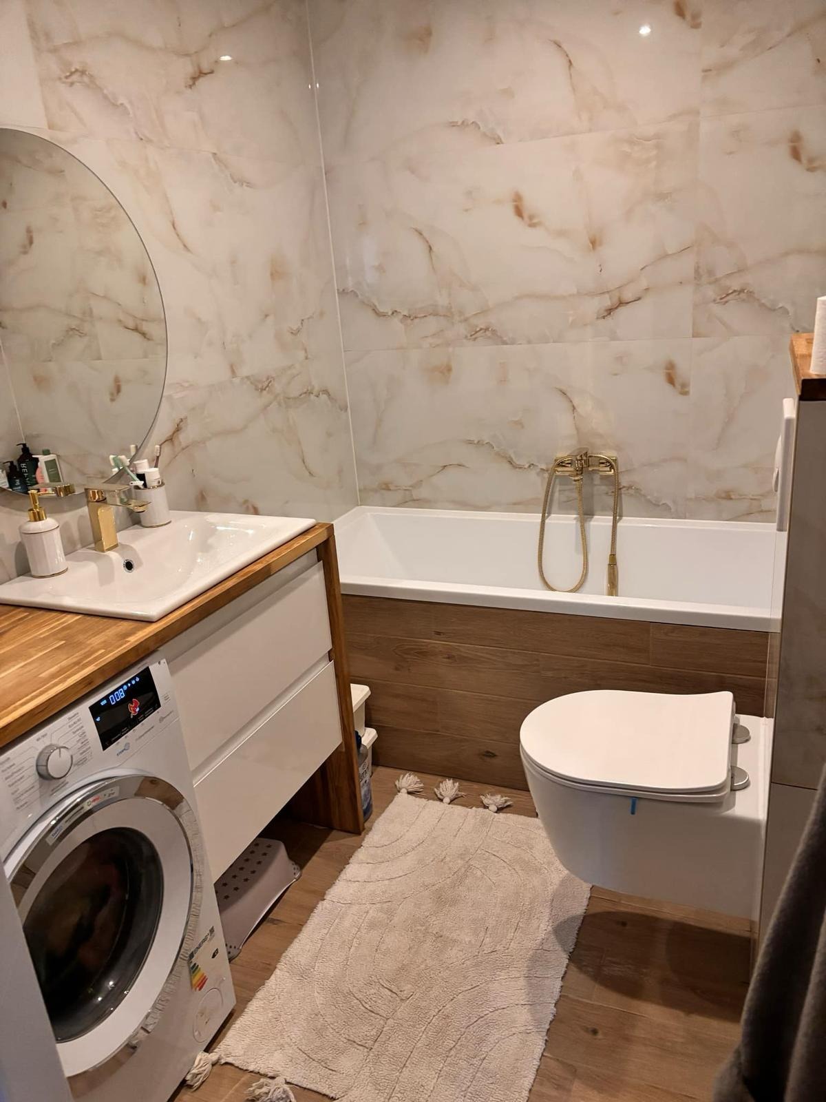 Nowoczesna łazienka z marmurowymi płytkami, drewnianymi akcentami i złotą armaturą, pralka z lewej strony.