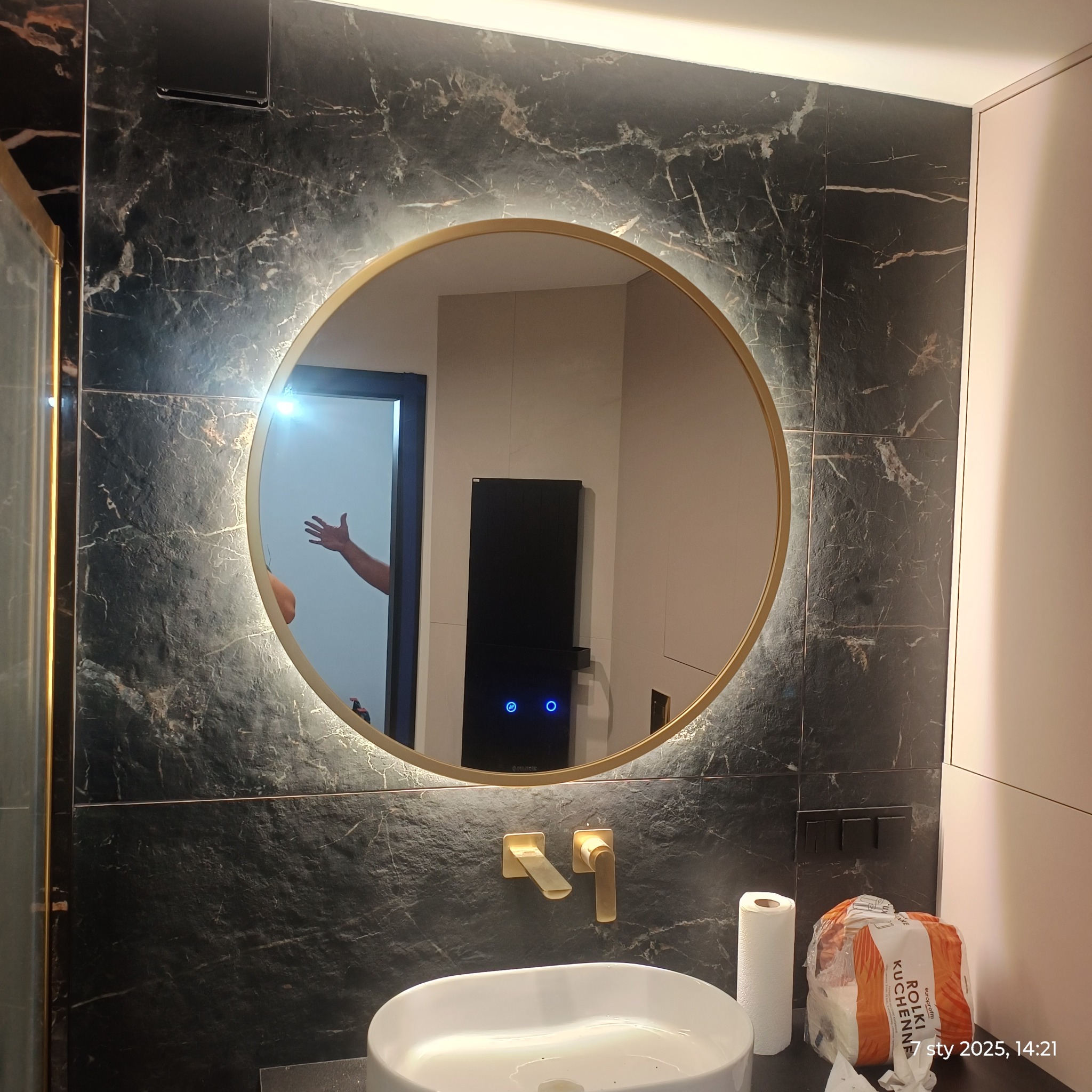 Łazienka z umywalką, okrągłym lustrem z podświetleniem LED, baterią w kolorze złotym i czarną marmurową ścianą z widocznym odbiciem osoby w lustrze