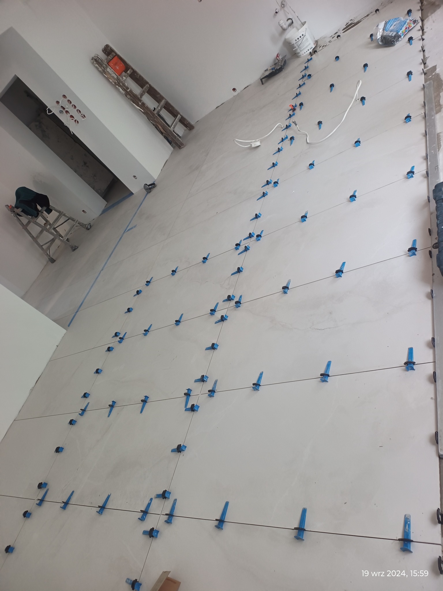 Świeżo ułożone, jasne płytki podłogowe z niebieskimi klipsami poziomującymi i czarnymi krzyżykami dystansowymi, widoczne drabiny i narzędzia w tle, w trakcie prac wykończeniowych.