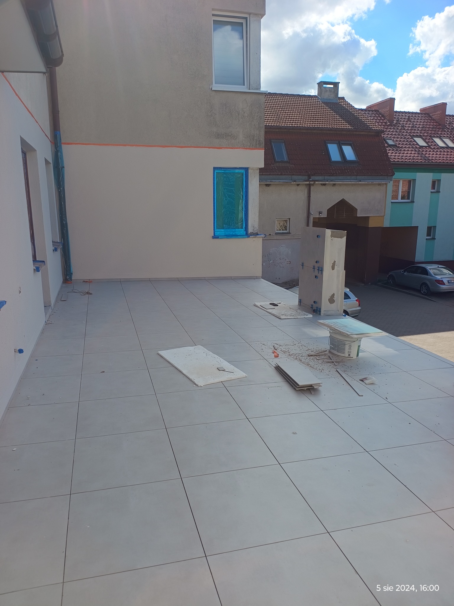 Układanie płytek na tarasie zewnętrznym budynku, widoczne narzędzia i resztki po cięciu płytek, okna zabezpieczone folią.