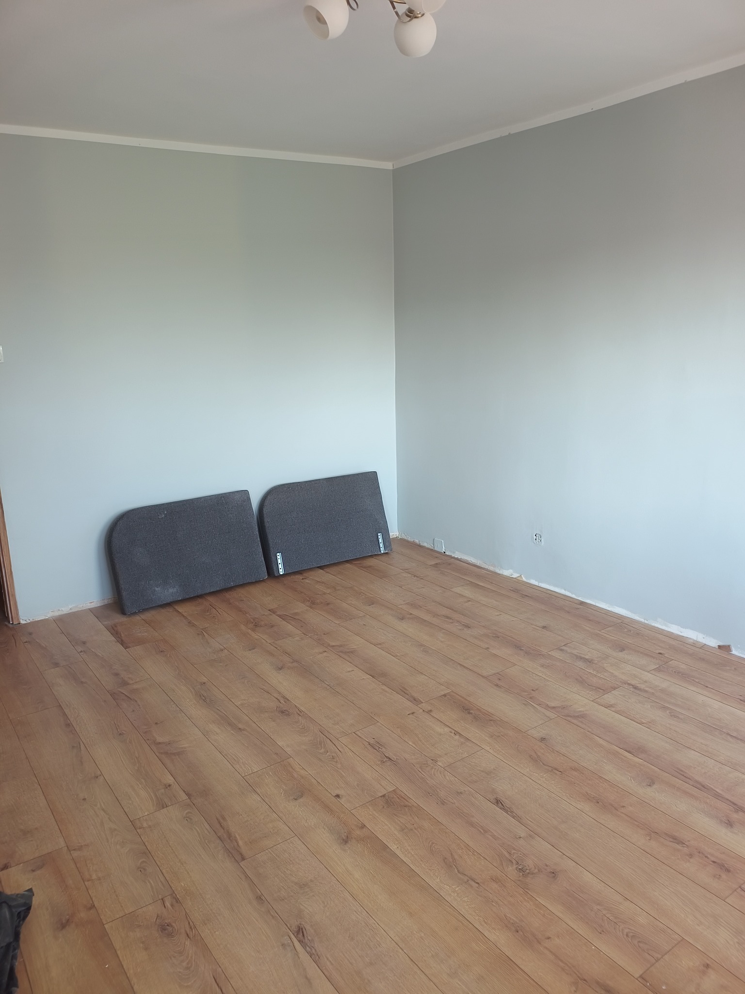 Pokój po remoncie: świeżo pomalowane szare ściany, ułożone panele podłogowe, przy ścianie oparte dwie płyty z szarego materiału.
