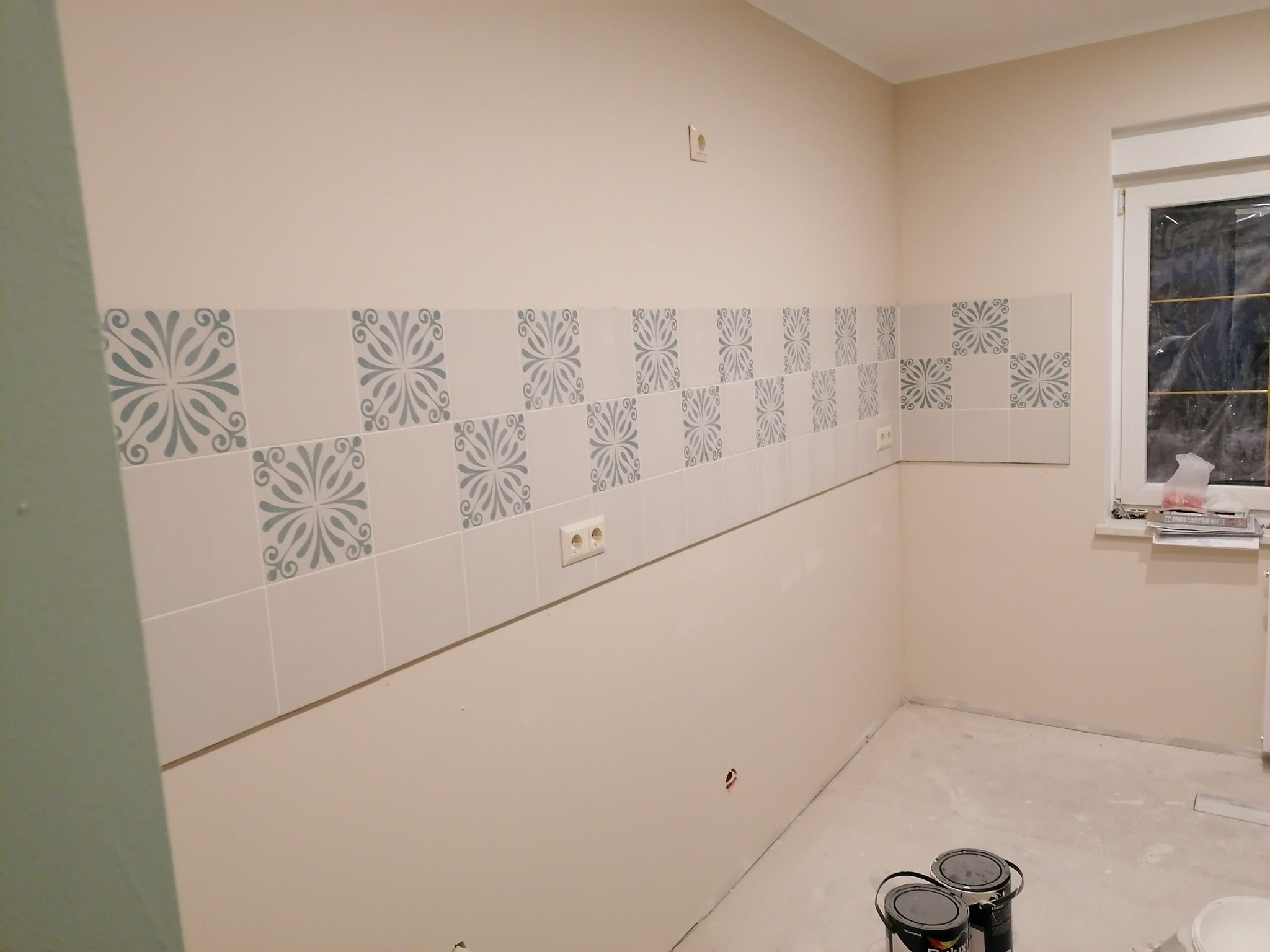 Ściana w trakcie remontu z nowo położonym pasem dekoracyjnych płytek ceramicznych z szarym wzorem, widoczne puszki z farbą i niedokończona podłoga.