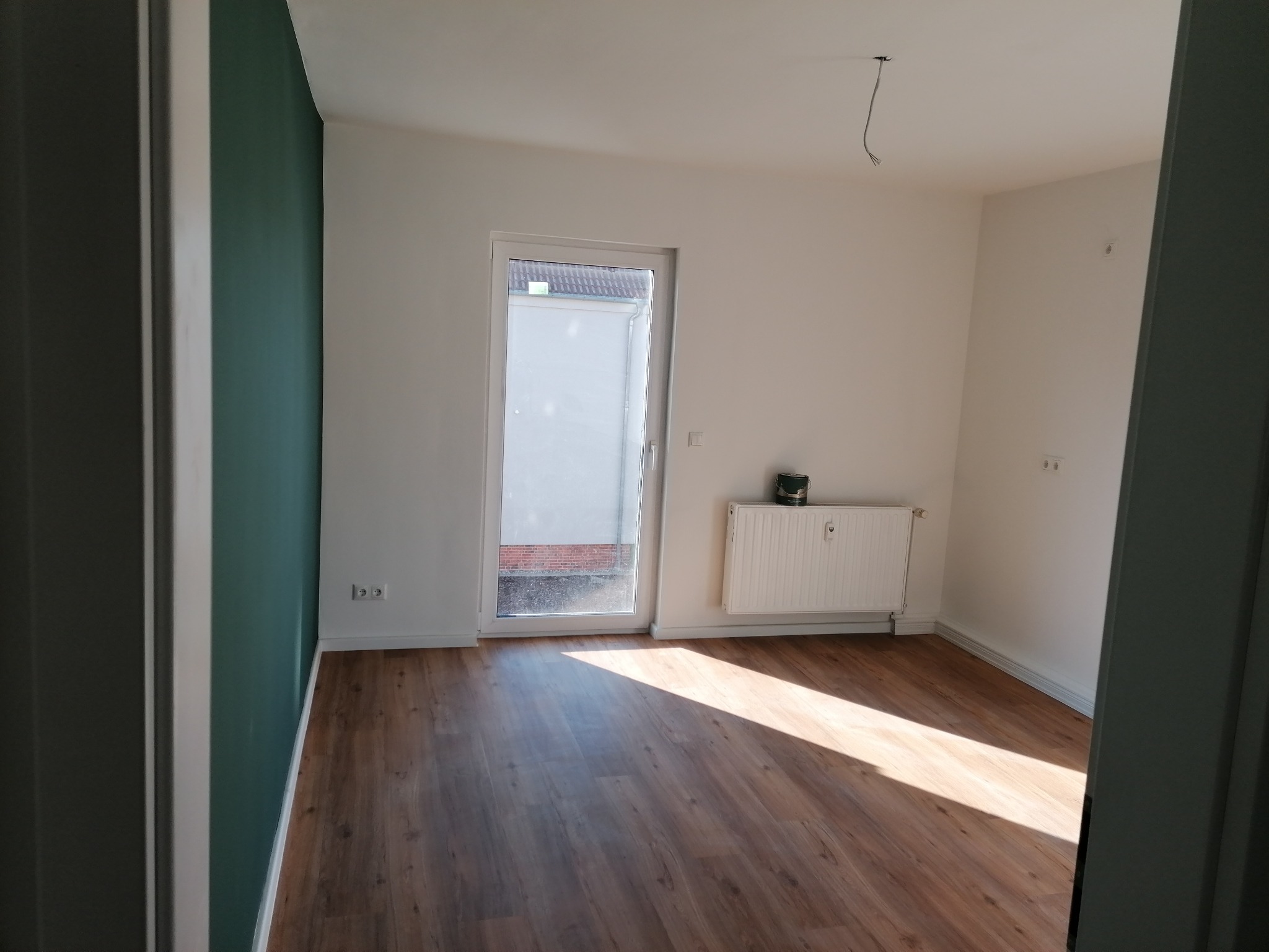 Pusty pokój po remoncie z panelami podłogowymi, pomalowaną na zielono ścianą i grzejnikiem pod oknem balkonowym, widoczne wystające kable ze ściany.