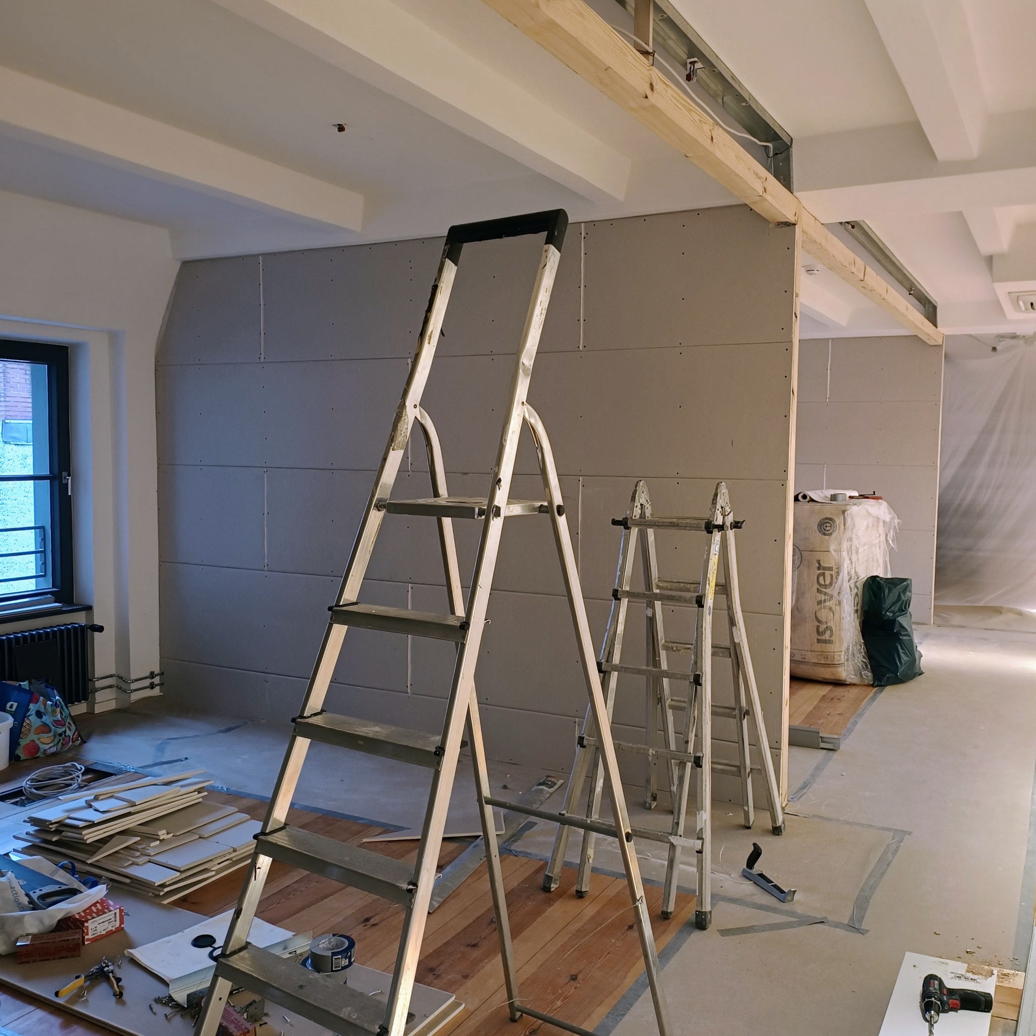 Wnętrze w trakcie prac wykończeniowych: zamontowane płyty gipsowo-kartonowe na ścianach, widoczne dwie drabiny aluminiowe, narzędzia pracy i materiały budowlane na podłodze zabezpieczonej folią...