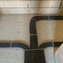 Układ czarnych, karbowanych rur instalacyjnych przytwierdzonych do surowej ściany za pomocą metalowych uchwytów. Rury rozchodzą się w trzech kierunkach, tworząc symetryczny wzór na tle szarego...